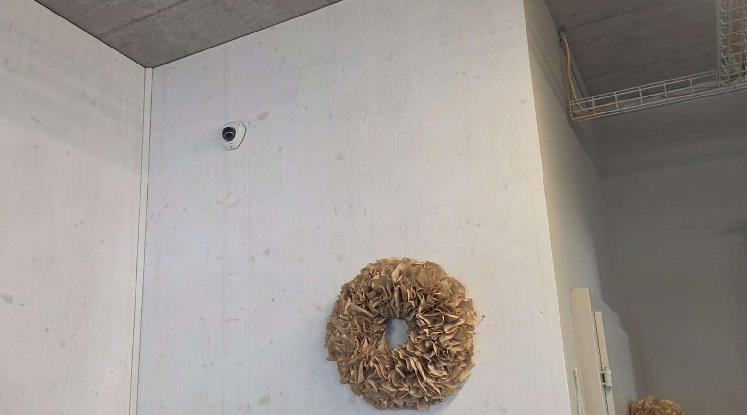 Kleine weiße Überwachungskamera dezent in der oberen Ecke einer hellen Holzwand im Blumenladen, daneben ein Deko-Kranz