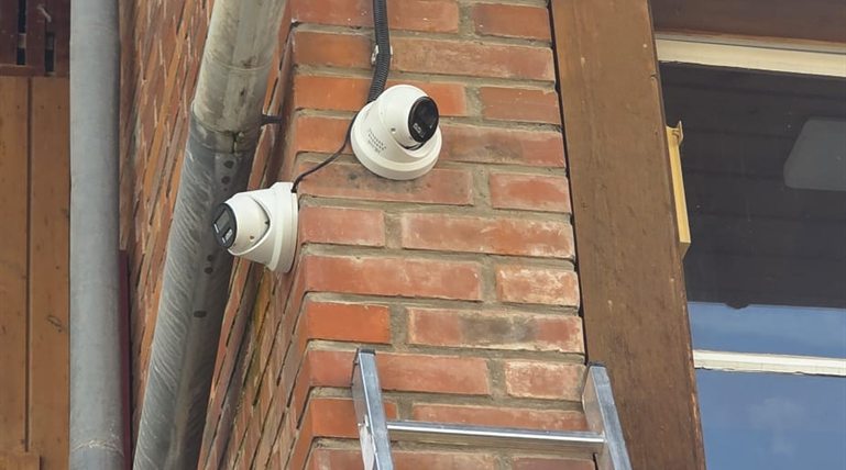 Zwei weiße Hikvision Dome-Kameras an einer Backstein-Hausecke unter Holz-Vordach, sichtbares Aufputz-Kabel