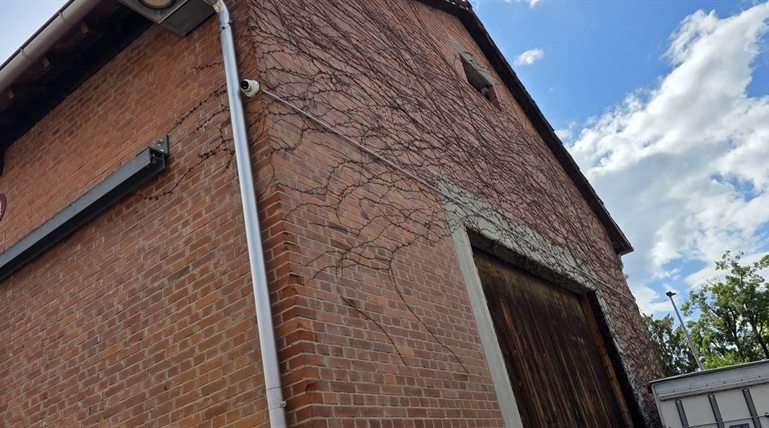Weiße Hikvision Dome-Kamera an der oberen Ecke eines Backstein-Giebels, Aufputz-Kabel entlang der Fassade