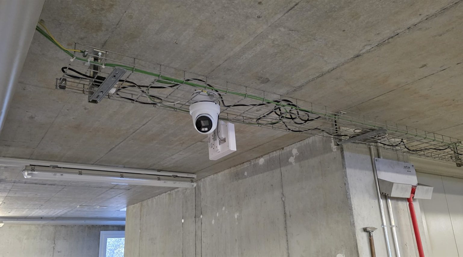 Weiße Hikvision ColorVu-Turret-Kamera an Halterung unter einer Betondecke, daneben eine Kabelpritsche mit Netzwerkkabeln im Lager