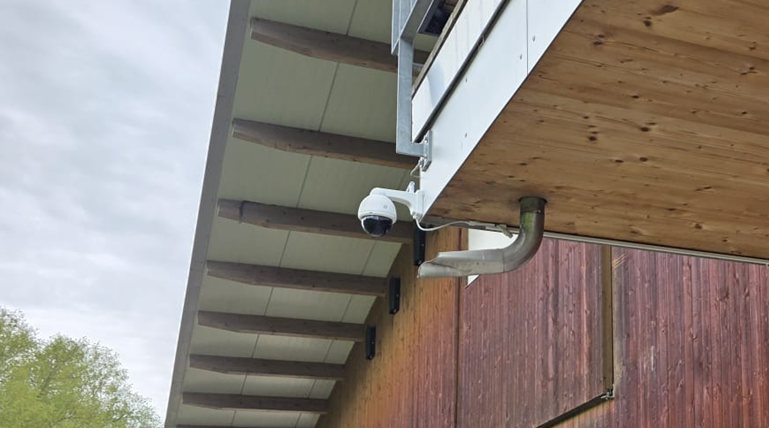 Nahaufnahme der weißen smart-cam Stall-Kamera (PTZ) an ihrer Metall-Halterung unter dem Vordach des Stalls
