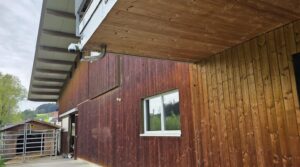 Weisse smart-cam PTZ-Stallkamera unter dem Dachvorsprung eines Holzstalls, im Hintergrund Weide mit Tierunterstand