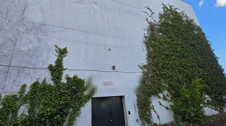Weiße, efeubewachsene Industriehalle mit einer Kamera über dem Anlieferungstor, Kabel sichtbar verlegt